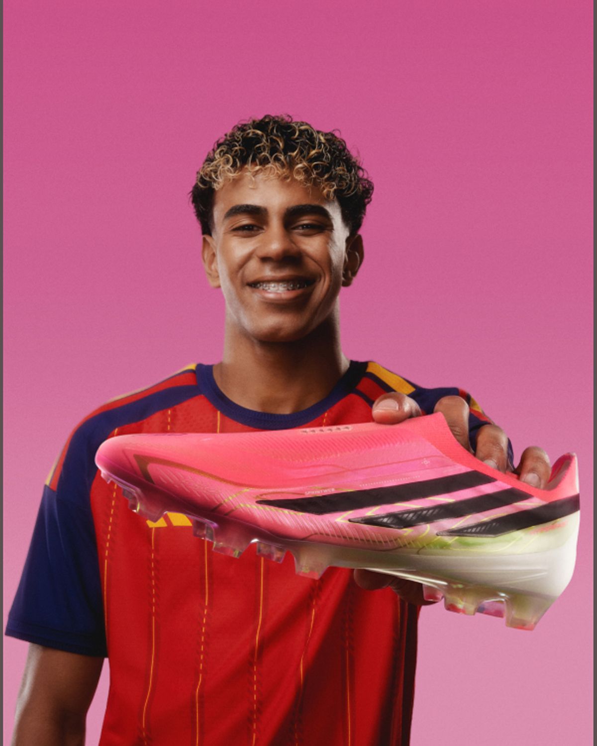 Smiling individual in a colorful sports jersey holds a pink and black soccer cleat against a gradient pink background.