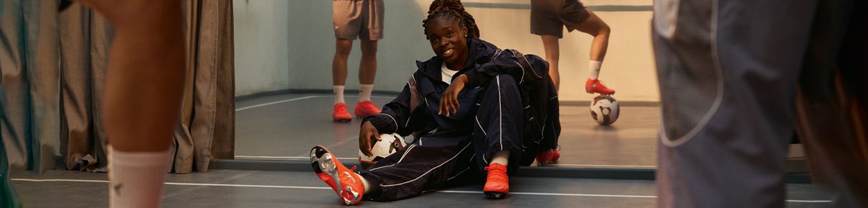 Person sitting on the floor in a soccer uniform, smiling, holding a ball, with teammates in the background, all wearing red soccer shoes.