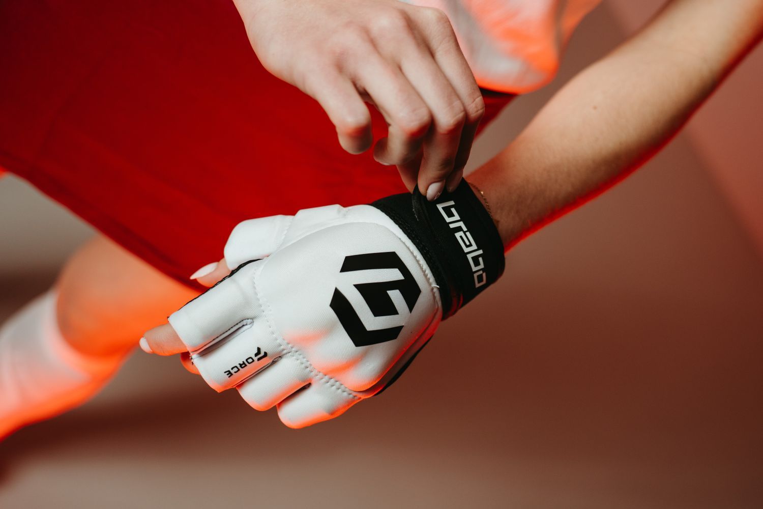 Close-up of a person adjusting a white Brabo hockey glove with black text and logo on the wrist, against a red background.