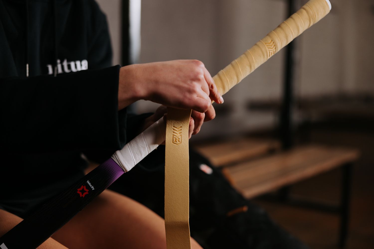Close-up of hands wrapping a hockey stick with yellow tape, wearing a black hoodie with text partially visible.