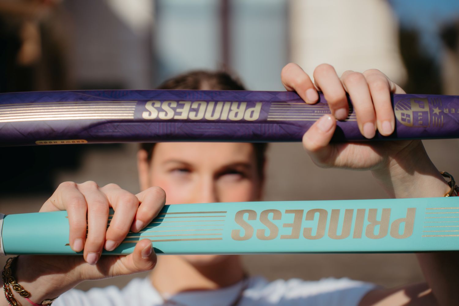 A person holds up two hockey sticks, one purple and one teal, both labeled "PRINCESS," with their face partially visible in the background.