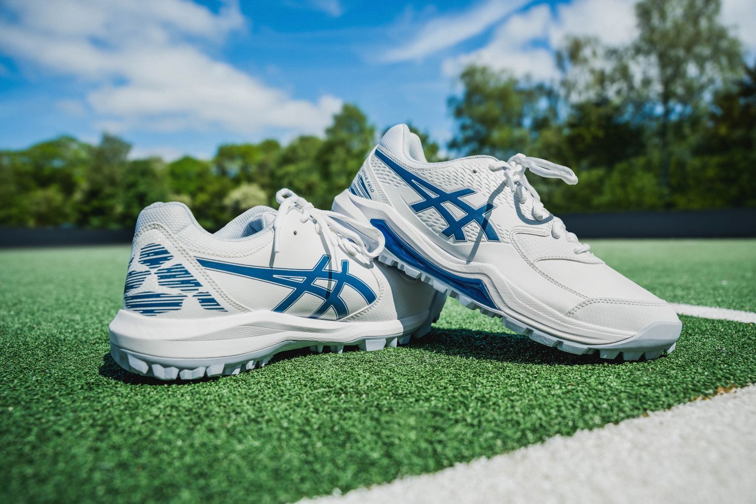 White and blue hockey shoes on green turf, with a blue sky and trees in the background.
