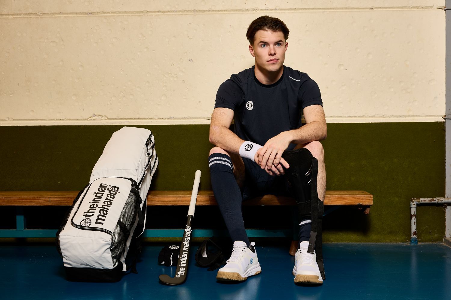 A hockey player sits on a bench in a locker room, wearing hockey gear and looking pensive, with a The Indian Maharadja hockey stick and bag beside him.