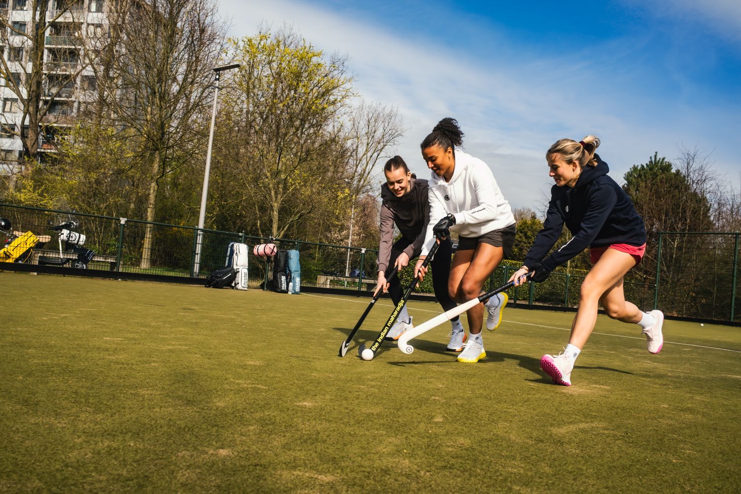 Three people playing field hockey on a green pitch, focused on the ball, with trees and sports bags in the background.