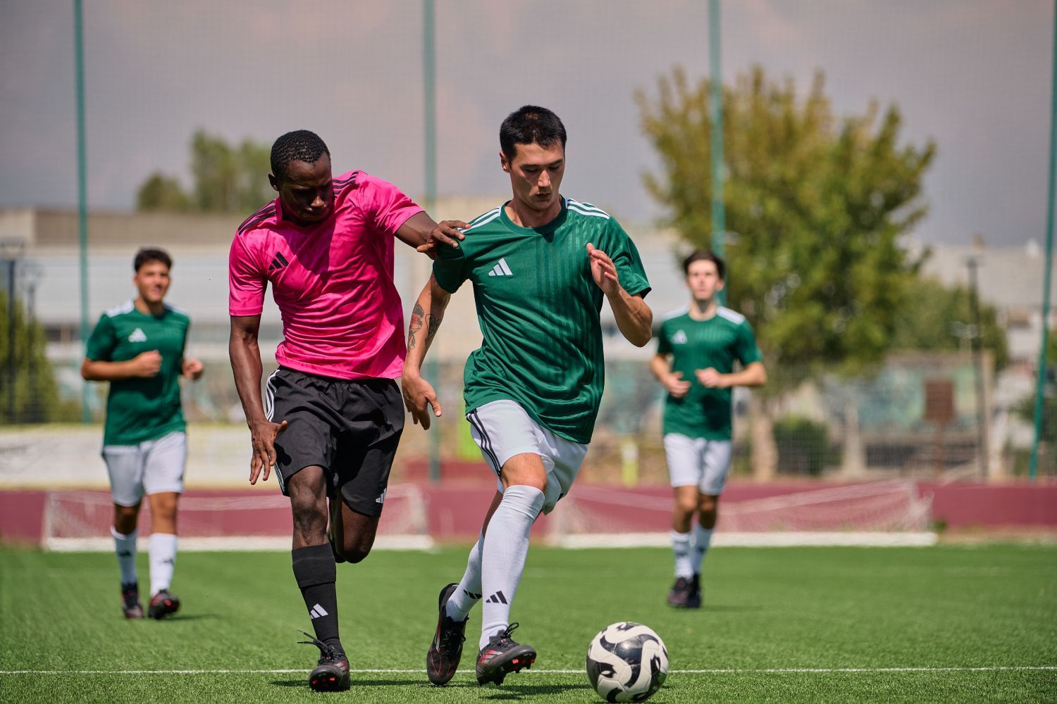 Hoe kies je de juiste voetbalsokken? Deskundig advies voor het veld Two soccer players, one in a pink jersey and the other in green adidas, compete for the ball on a sunny field, with teammates and a goal in the background.