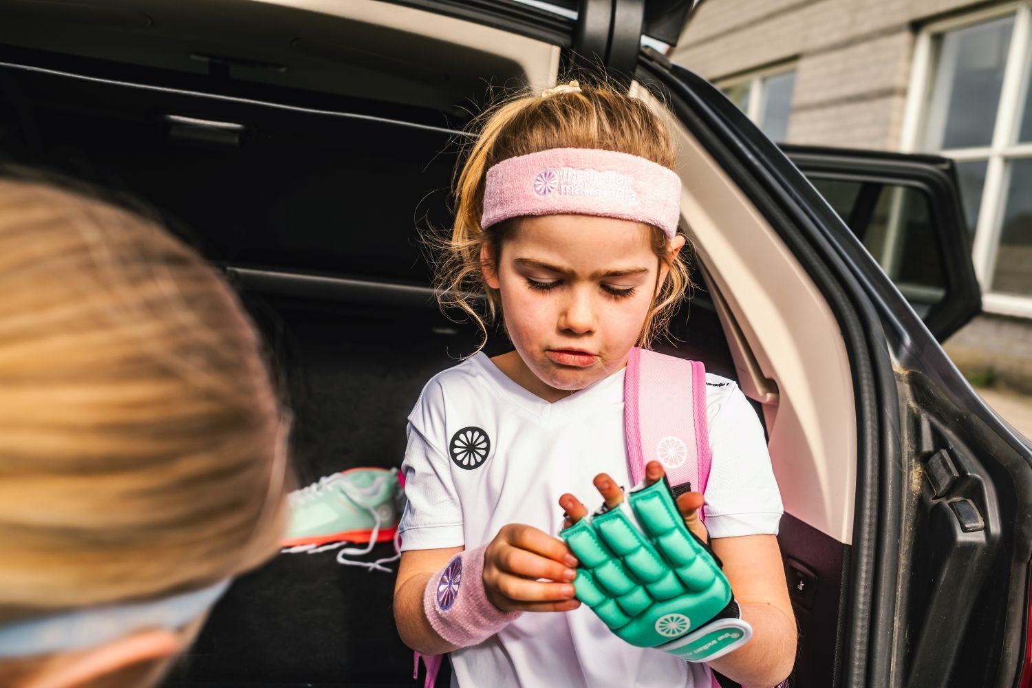 Young girl in a white shirt and pink headband puts on a padded hockey glove, standing by a car trunk with sports gear.