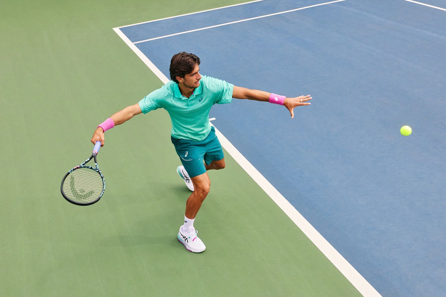 Tennis player in green outfit lunges to hit a yellow ball with a racket on a blue and green court.