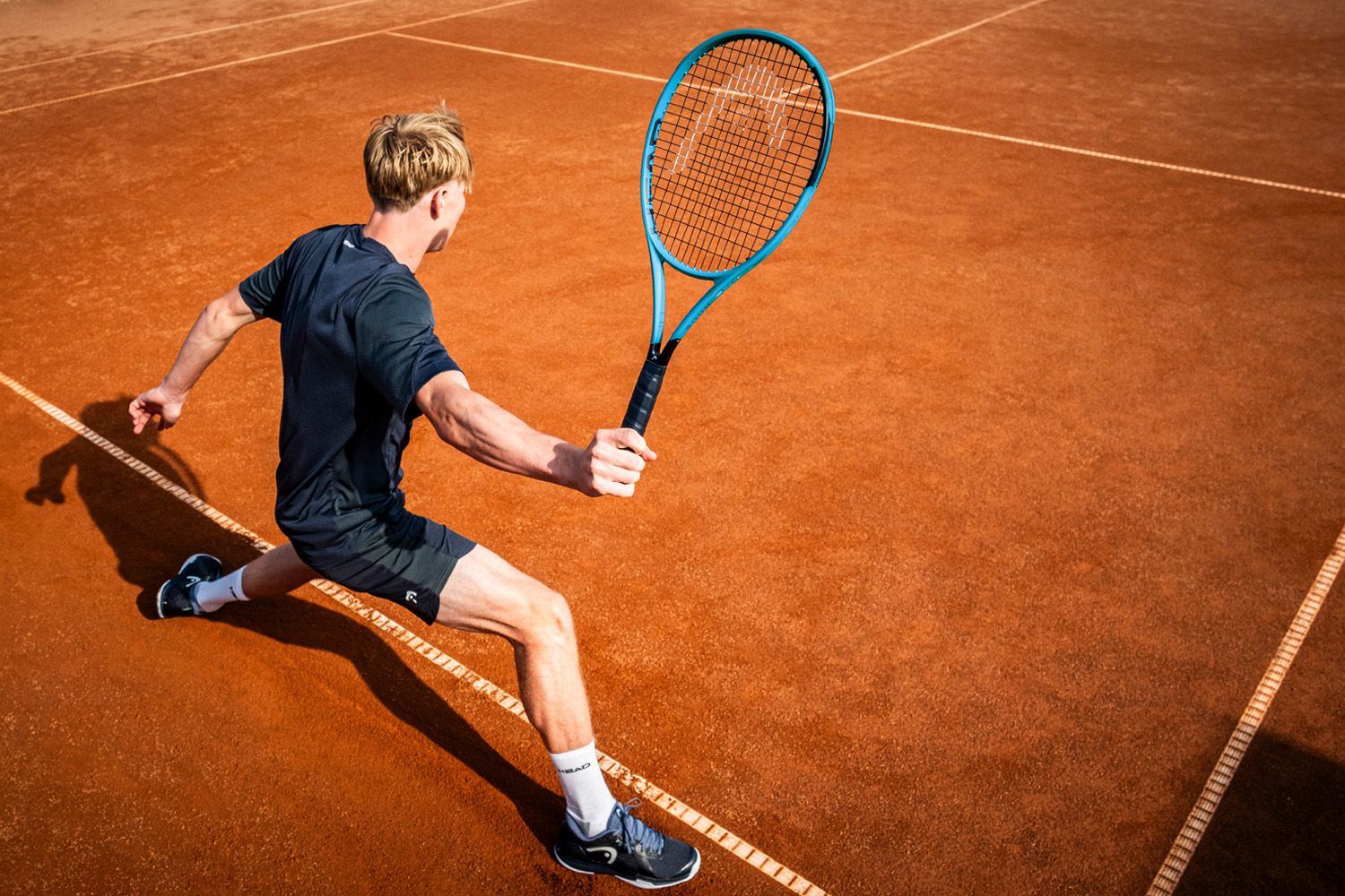 Tennis player in a black outfit lunging to hit a backhand shot on a clay court.