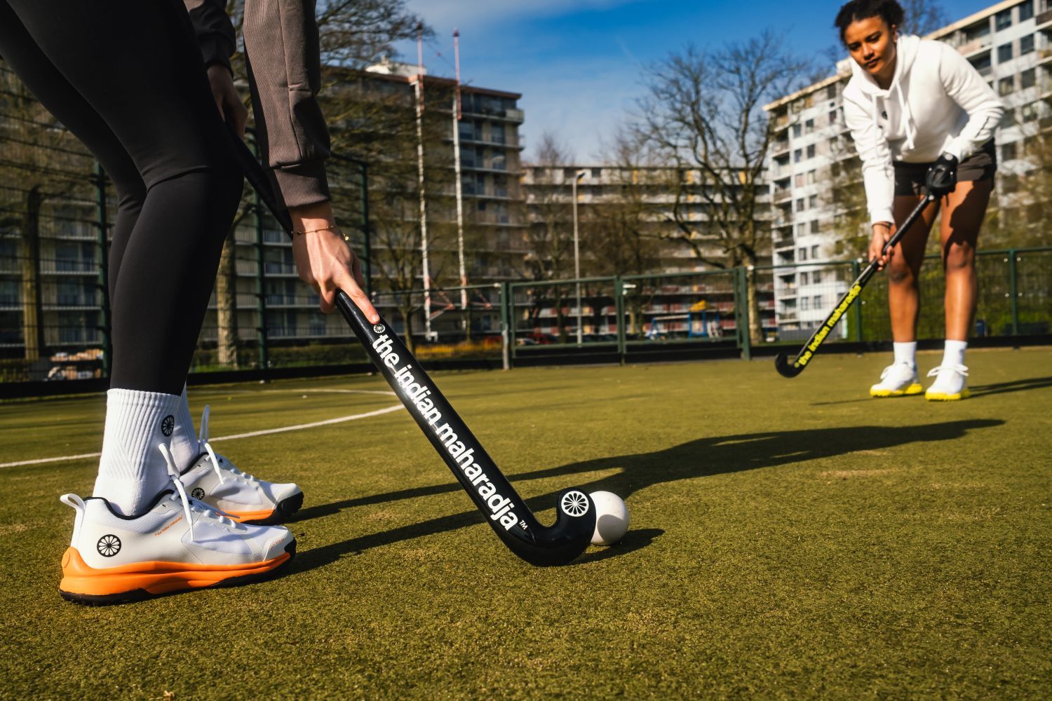 Two people playing field hockey on a sunny day, with one about to hit the ball. Apartments and trees are visible in the background.