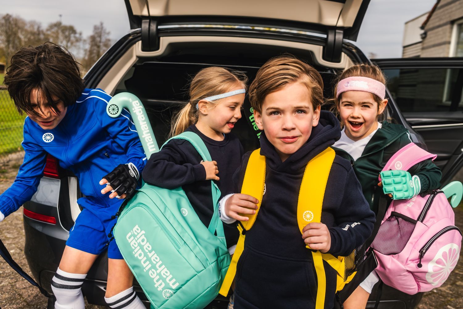 Children with hockey gear and colorful hockeybags from The Indian Maharadja emerge from a car trunk, smiling and ready for an outdoor activity.