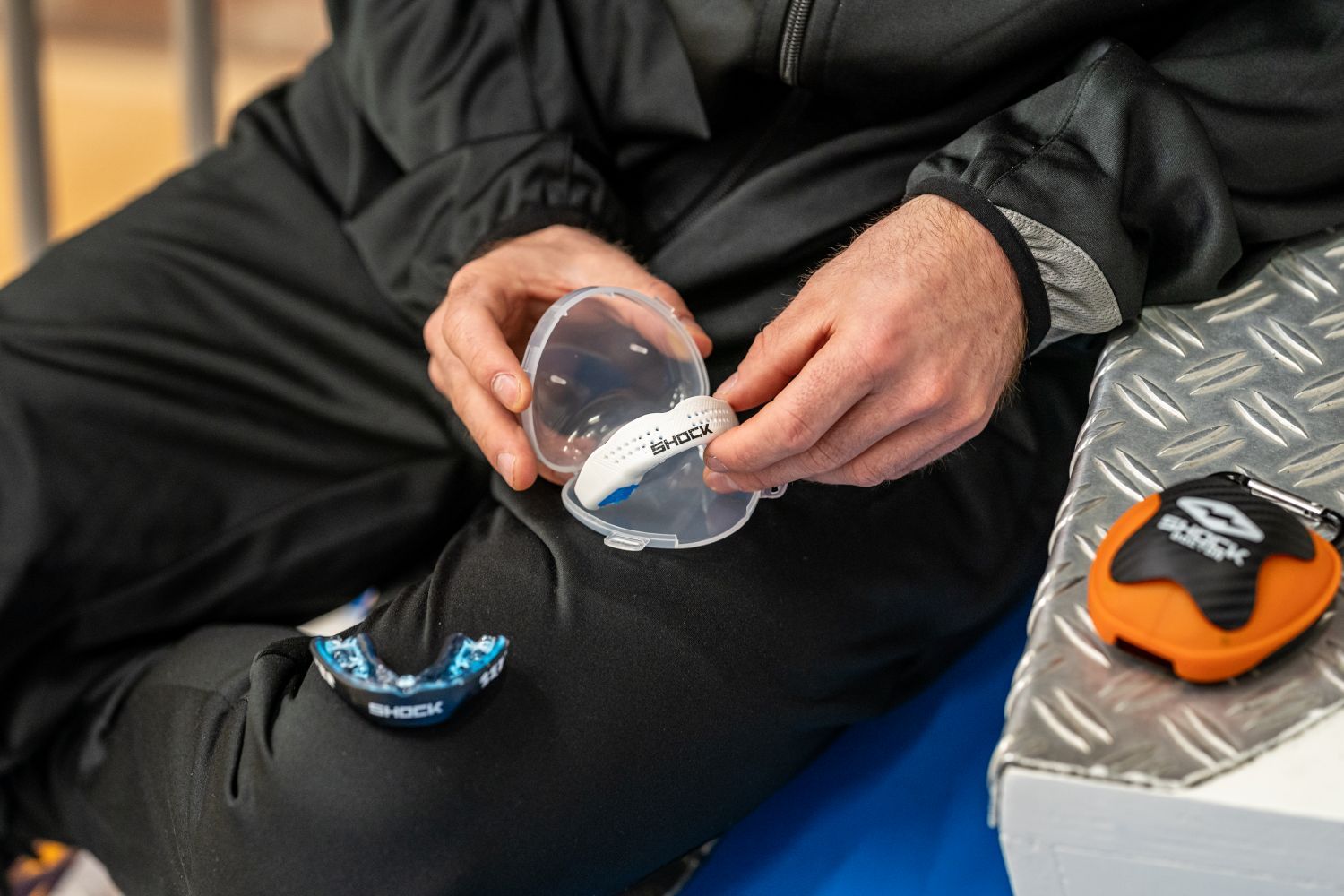 Hoe kies je het beste hockey bitje? Person in black clothing holding a hockey mouthguard in a clear case, with additional shockdoctor mouthguards and cases nearby.