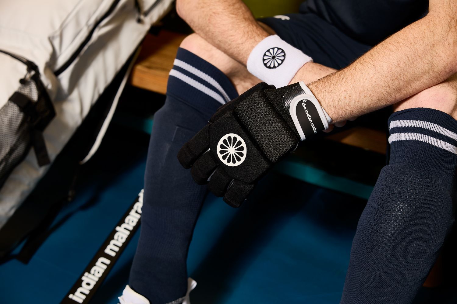 A person wearing black hockey gloves, navy shorts, and socks sits on a bench. A hockey stick and a bag are nearby.