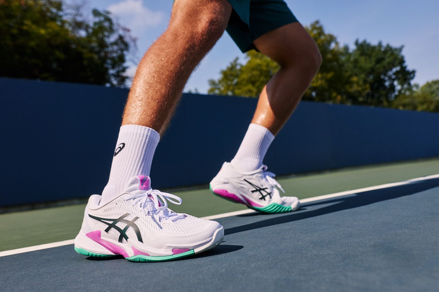 Person wearing white Asics sneakers with green and pink accents on a tennis court, showing mid-stride motion.