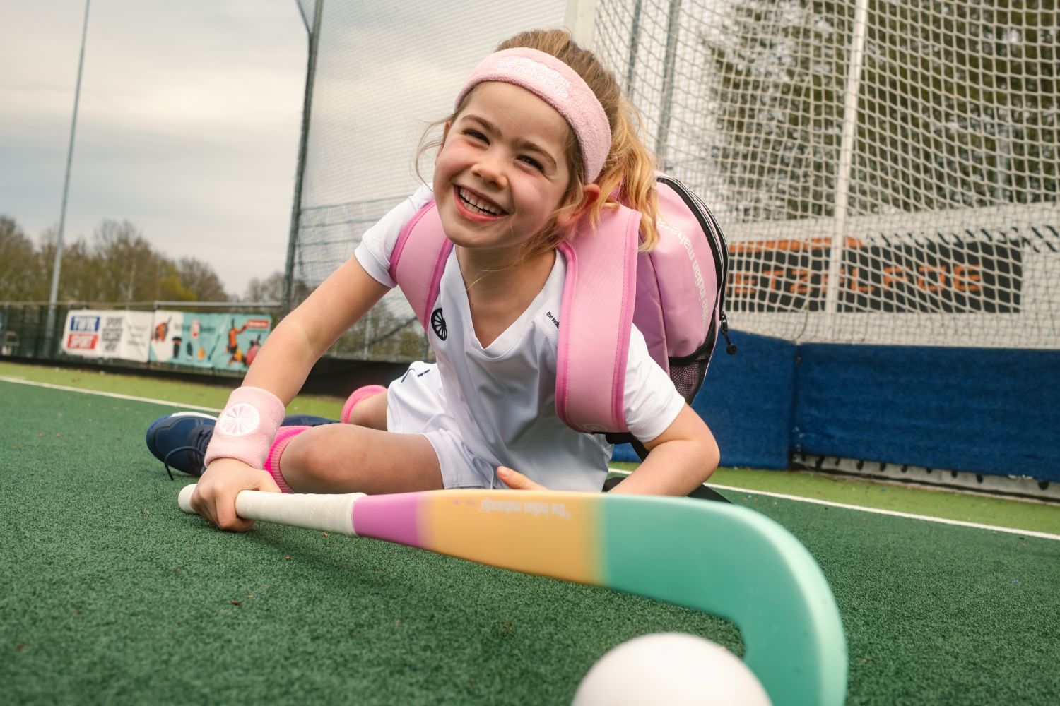 Smiling girl with a hockey stick and pink backpack lying on a field near a goal net, wearing a white hockey outfit and headband.