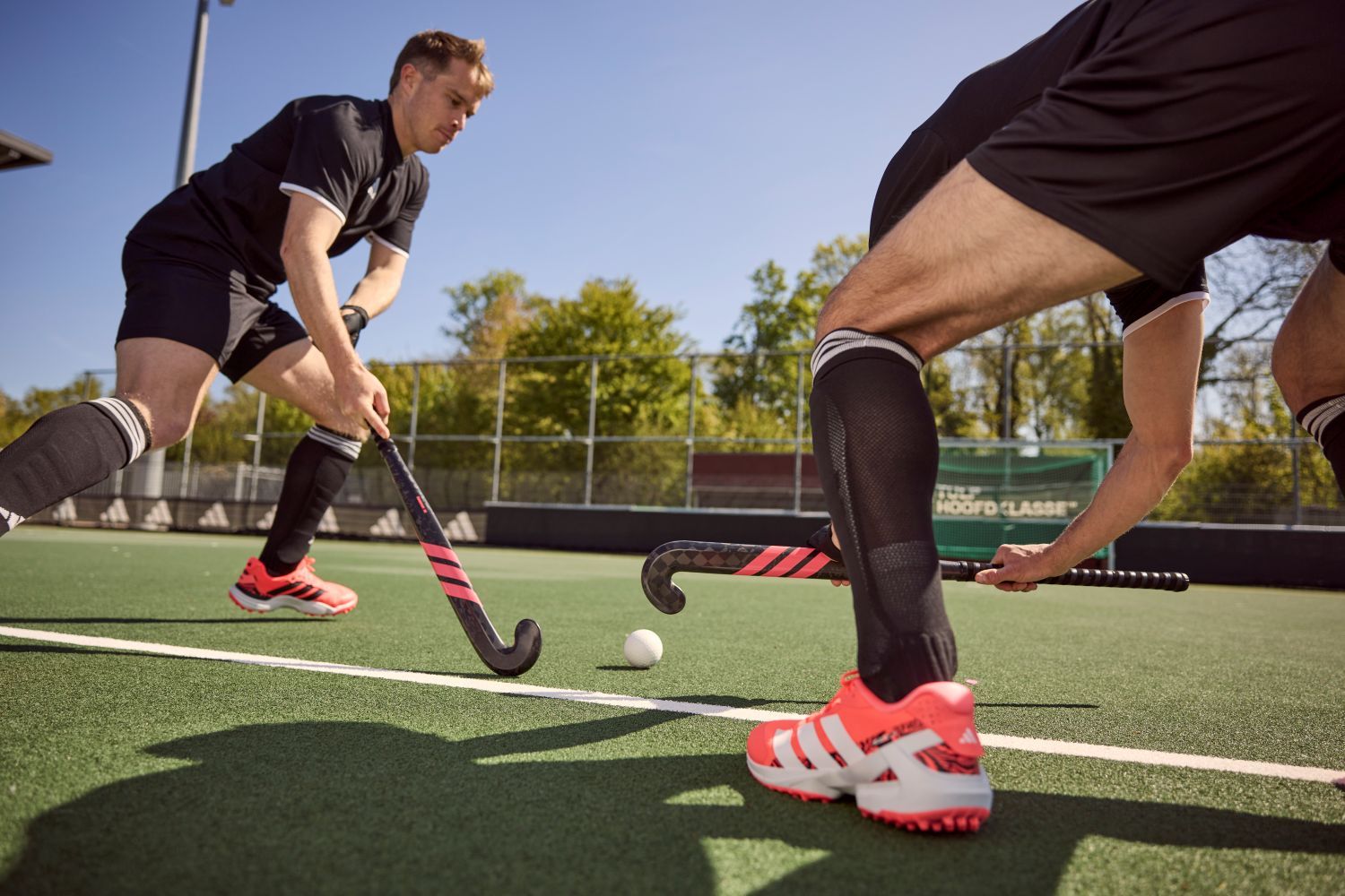 Hockeytermen van A tot Z: Het ultieme hockeywoordenboek Two field hockey players in black uniforms and bright orange shoes competing for the ball on a sunny outdoor field.