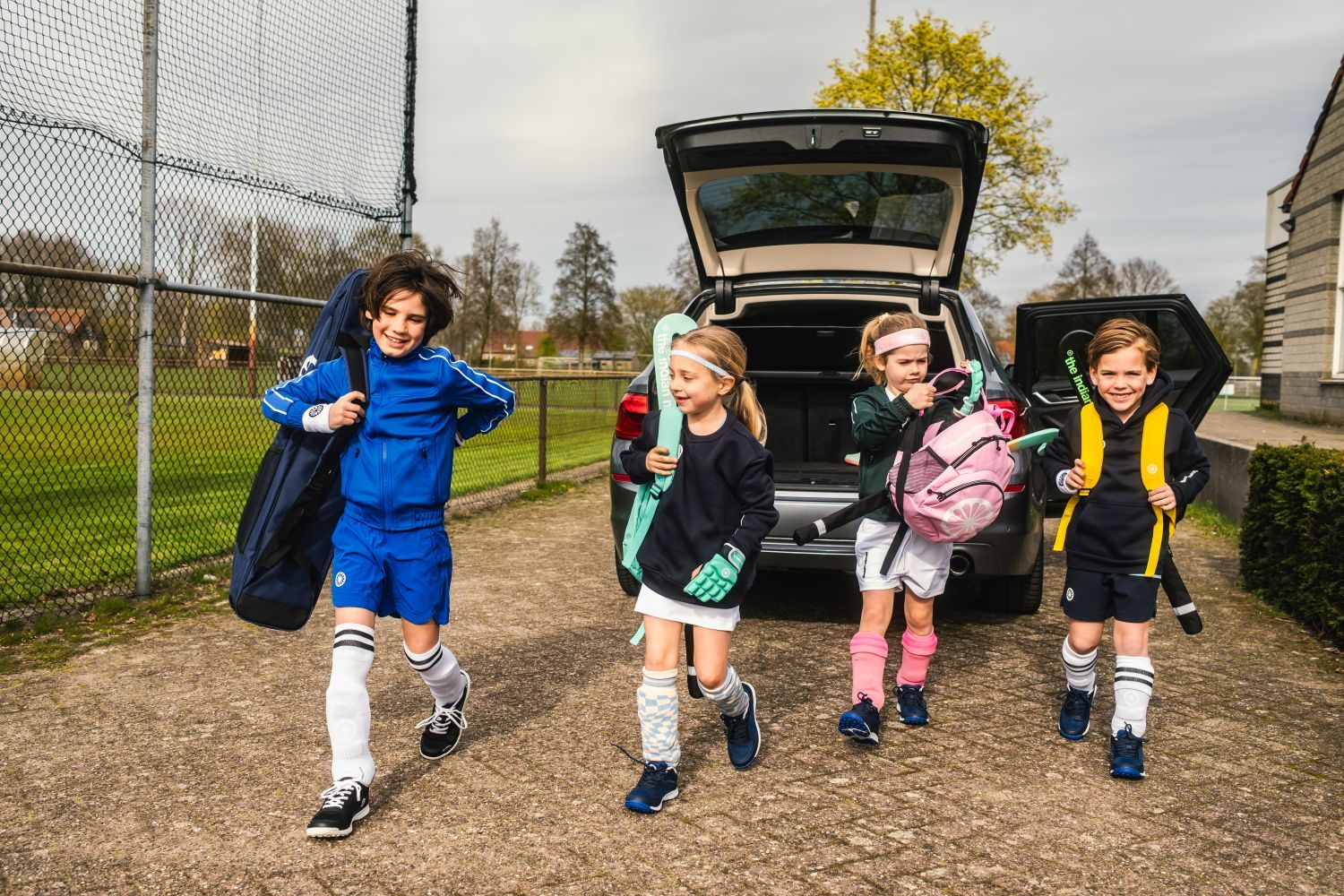 Welke hockeyspullen heb je nodig? Alles voor je eerste training Four children in The Indian Maharadja hockey gear excitedly walk away from an open SUV, carrying hockey sticks and bags, next to a fenced field.