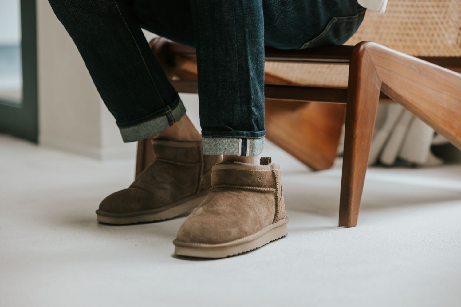Person in cuffed jeans sitting on a chair, wearing brown suede boots on a light-colored floor.