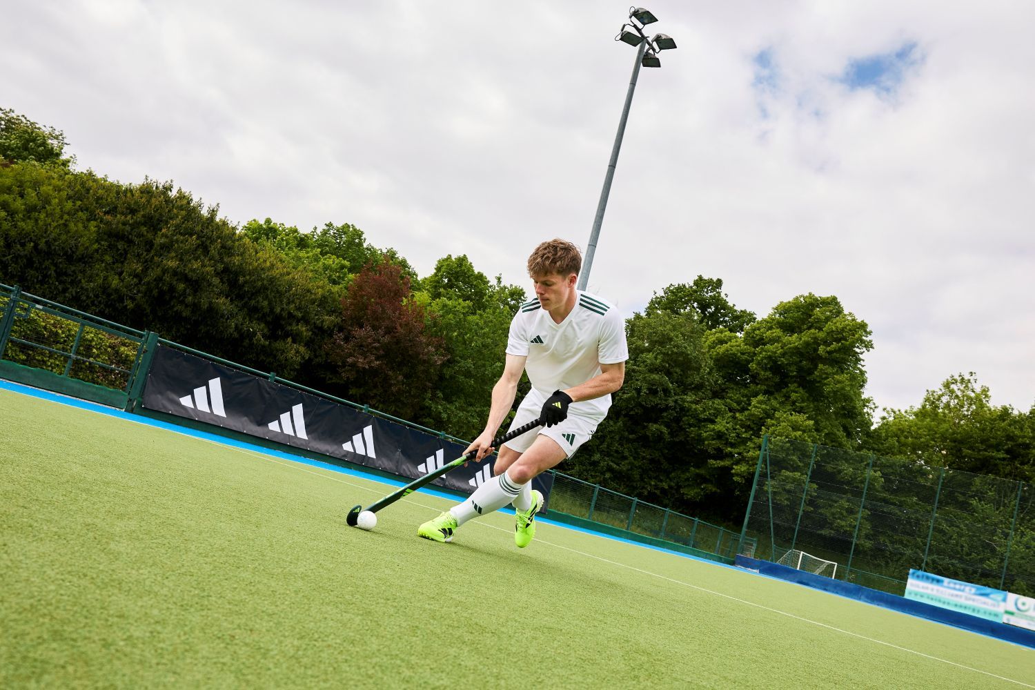 Welke adidas hockeystick past bij mij? De ultieme gids A field hockey player in a white uniform and neon green shoes skillfully maneuvers with the ball on a green pitch, surrounded by trees and a fenced boundary.