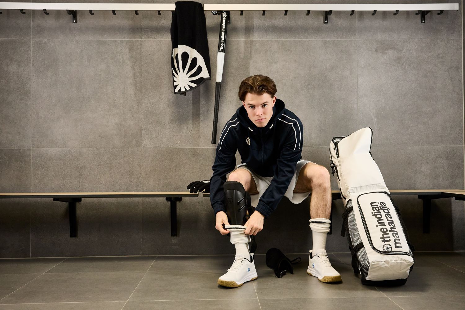 Welke scheenbeschermers heb je nodig voor hockey? A person in The Indian Maharadja hockey attire sits on a bench in a locker room, adjusting shin guards, with equipment, a sports bag, and a towel visible nearby.