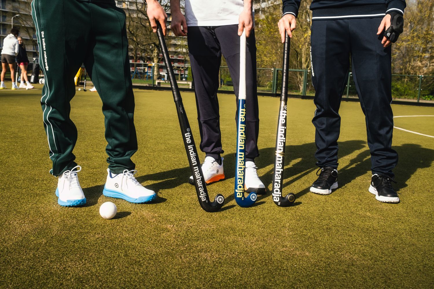 Hoe kies je de juiste maat hockeystick? Vind de perfecte lengte Three field hockey players stand on the turf with sticks labeled "The Indian Maharadja" and a ball in front of them.
