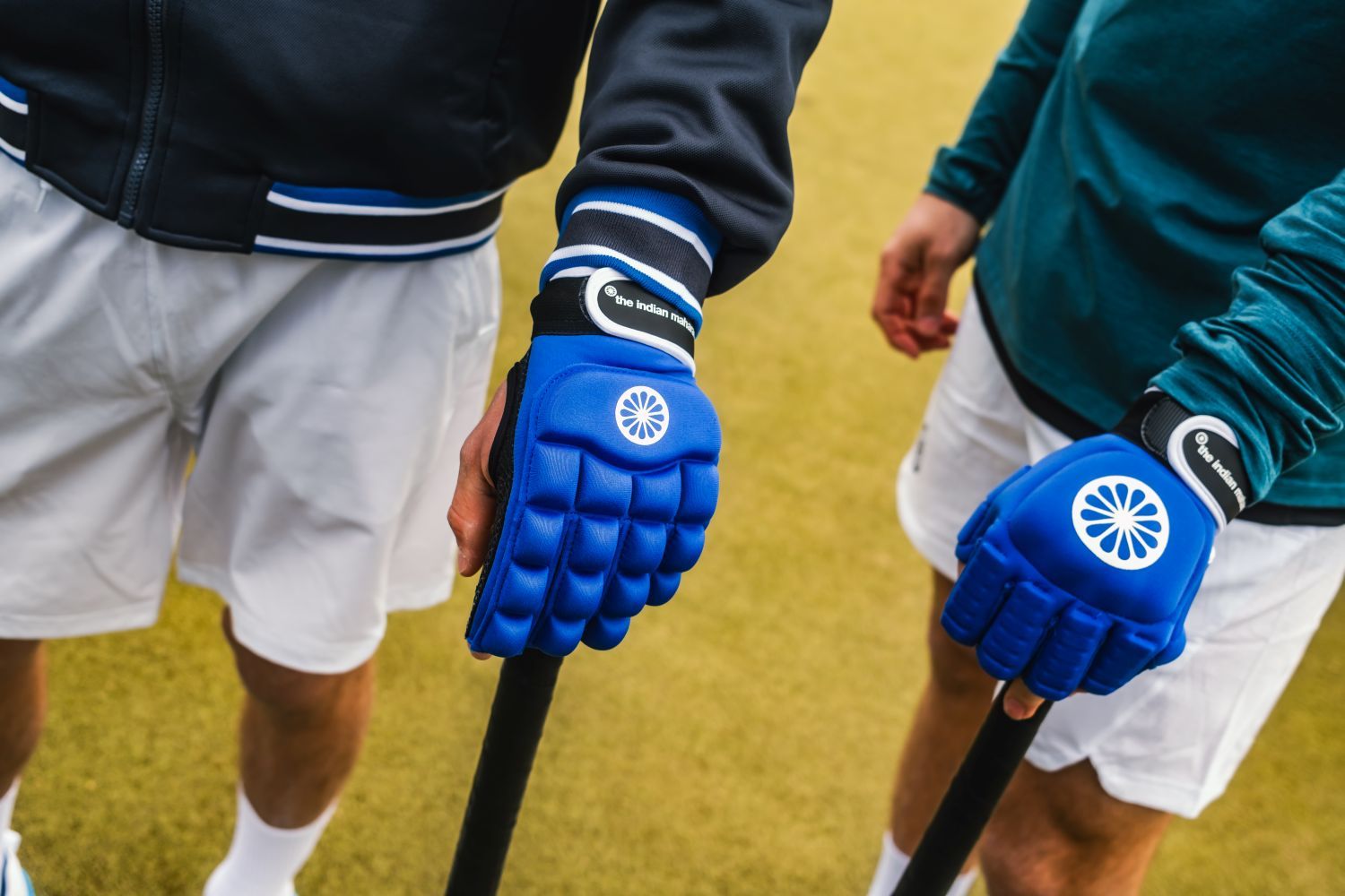 Hockey handschoen kiezen: Hoe vind je de beste bescherming? Two people holding hockey sticks, wearing blue padded gloves on a grass field. Both are in sports attire.