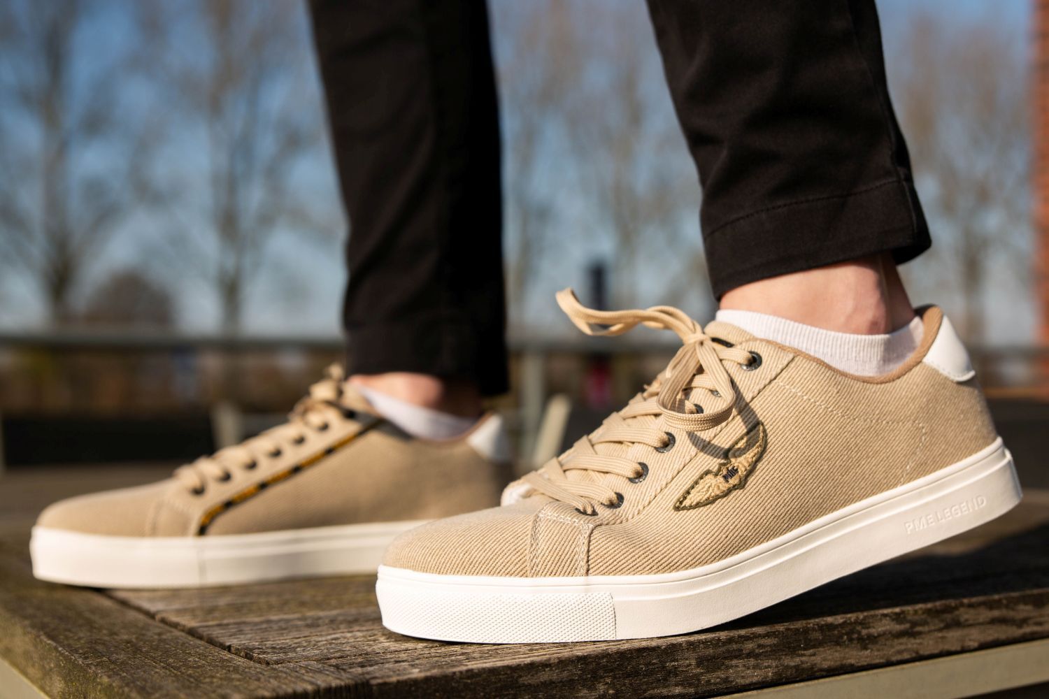 Person wearing beige sneakers with white soles and black pants stands on wooden surface outdoors.