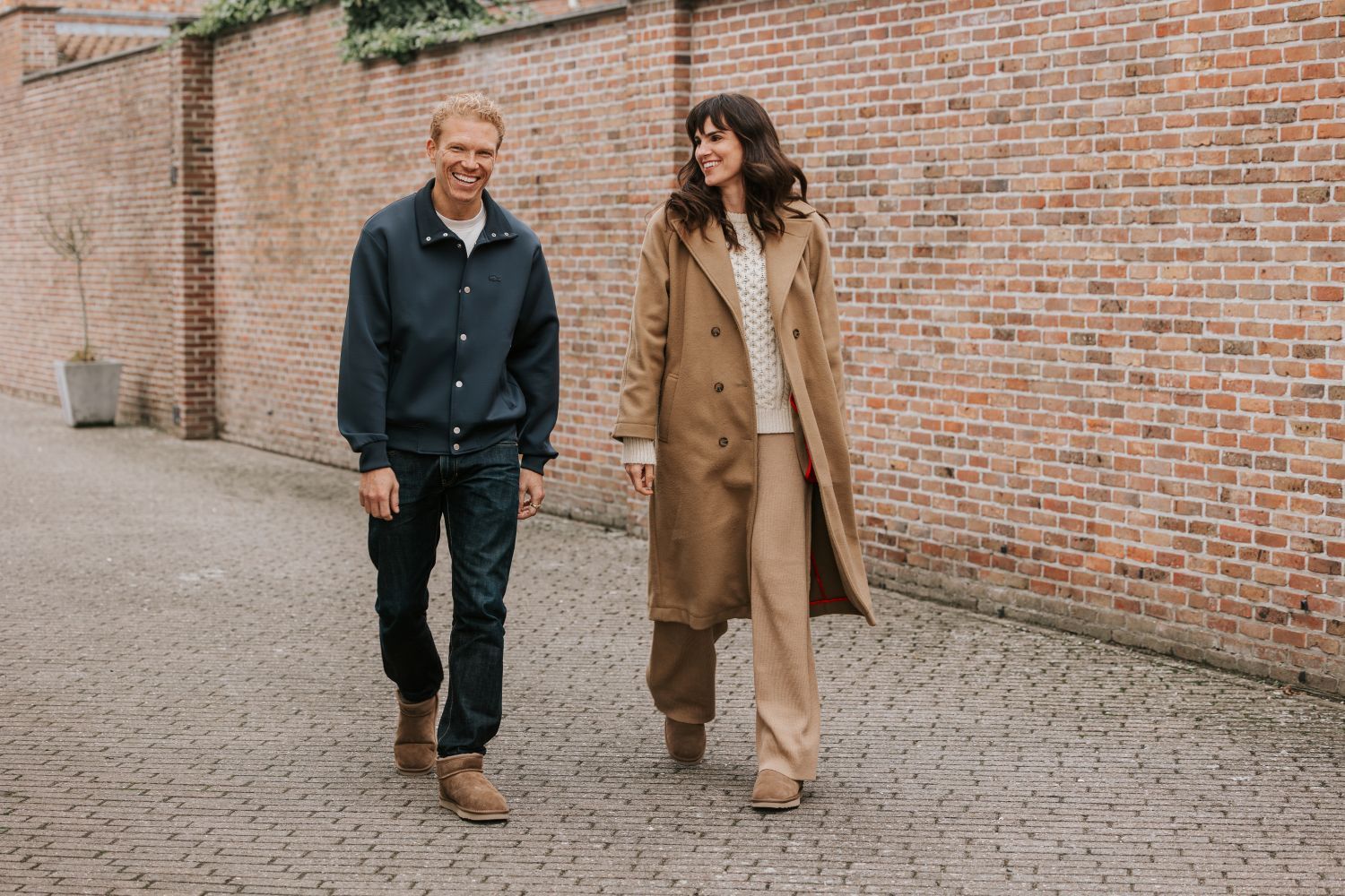 A man and a woman walking outdoors, smiling, dressed in casual winter clothing with a brick wall in the background.