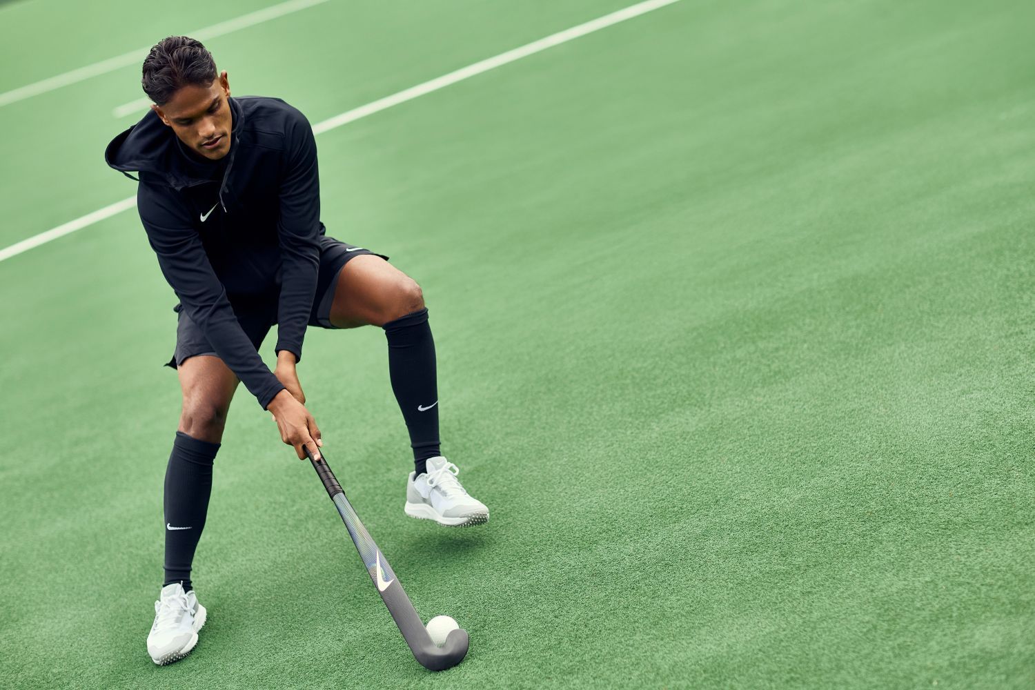 A field hockey player in black Nike sportswear and white shoes practices on a green turf field, focusing on hitting a white ball with a hockey stick.