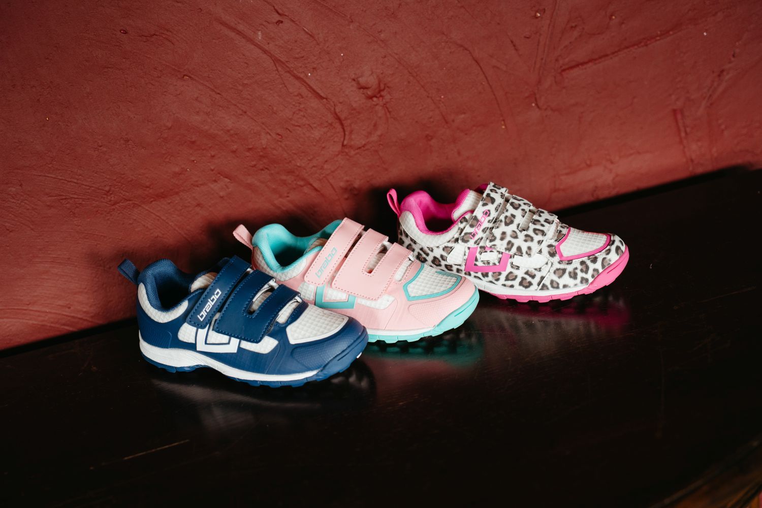 Three kids' Brabo hockey shoes with velcro straps displayed: blue, pink, and leopard print, on a dark surface against a red wall.