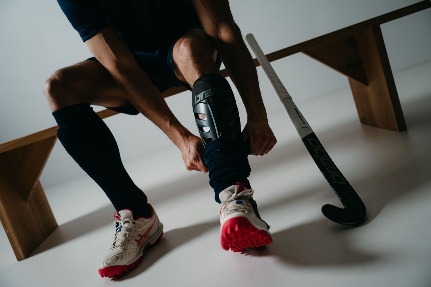 A person sitting on a bench adjusts a Brabo shin guard under a sock, with a field hockey stick resting nearby on the floor.
