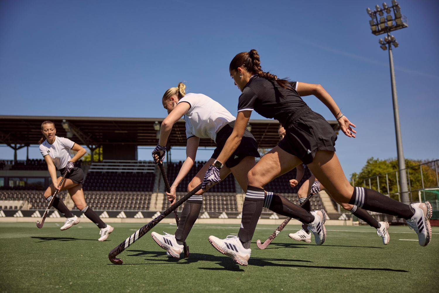 Hoe kies je het juiste materiaal van een hockeystick? Women playing field hockey on a sunny day, wearing white and black uniforms, running with sticks on a green field with bleachers in the background.