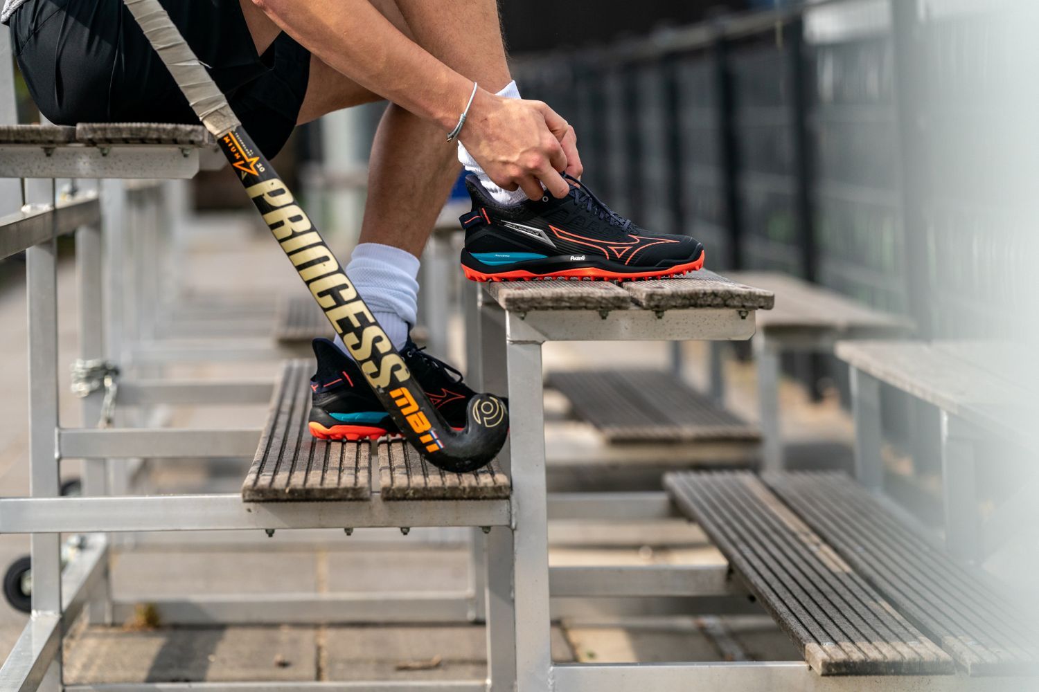 Het verschil tussen dames hockeyschoenen en heren hockeyschoenen Person tying black Mizuno hockey shoes, holding a hockey stick on metal bleachers.