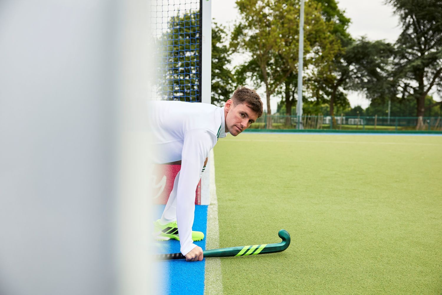 Man in white sports attire crouches with a adidas ChaosFury hockey stick near a field goal, looking towards the camera. Trees and a fence are in the background.