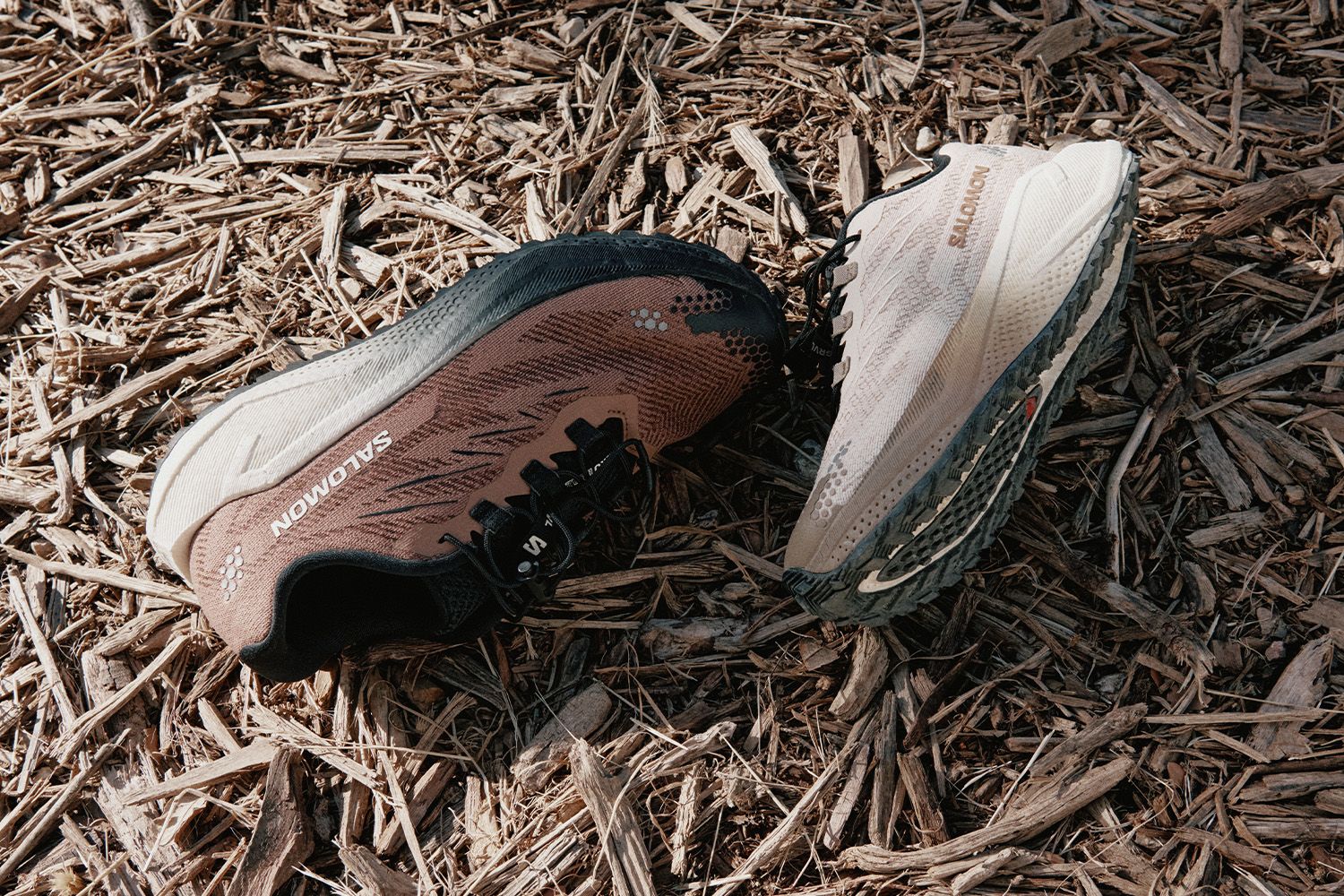 Two running shoes on a bed of wood chips, one brown and black, the other beige and gray, with visible brand logos.