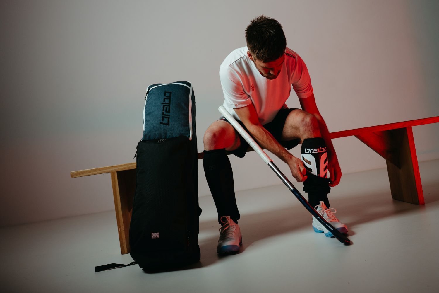 Wat is het verschil tussen zaal- en velduitrusting voor hockey? A person in Brabo hockey gear adjusts knee pads, seated on a bench with a hockey stick and a bag beside them in a dimly lit room.