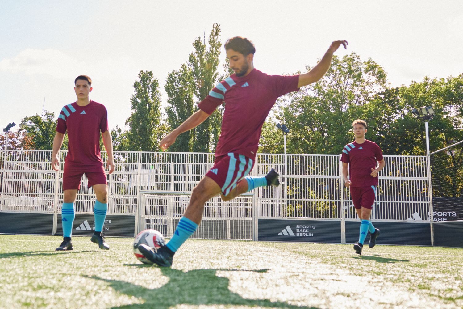 Three soccer players in maroon and blue adidas kits on a sunny field, one kicking a ball, with trees and a metal fence in the background.