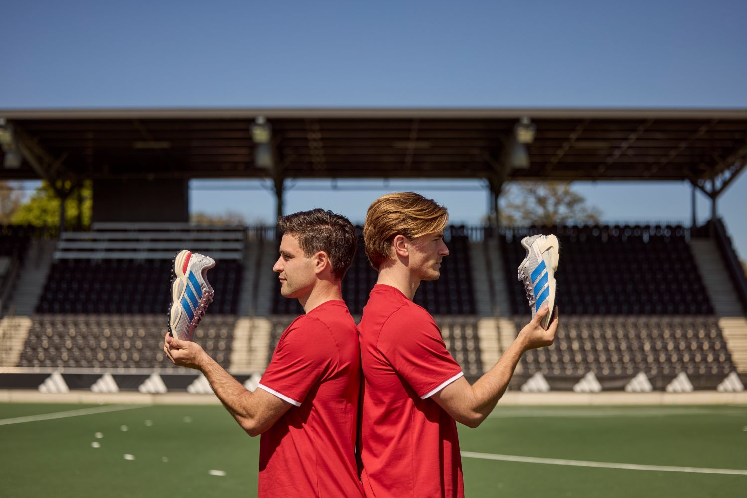 Two men in red shirts stand back-to-back on a sports field, each holding a adidas shoe with blue stripes, facing empty bleachers.