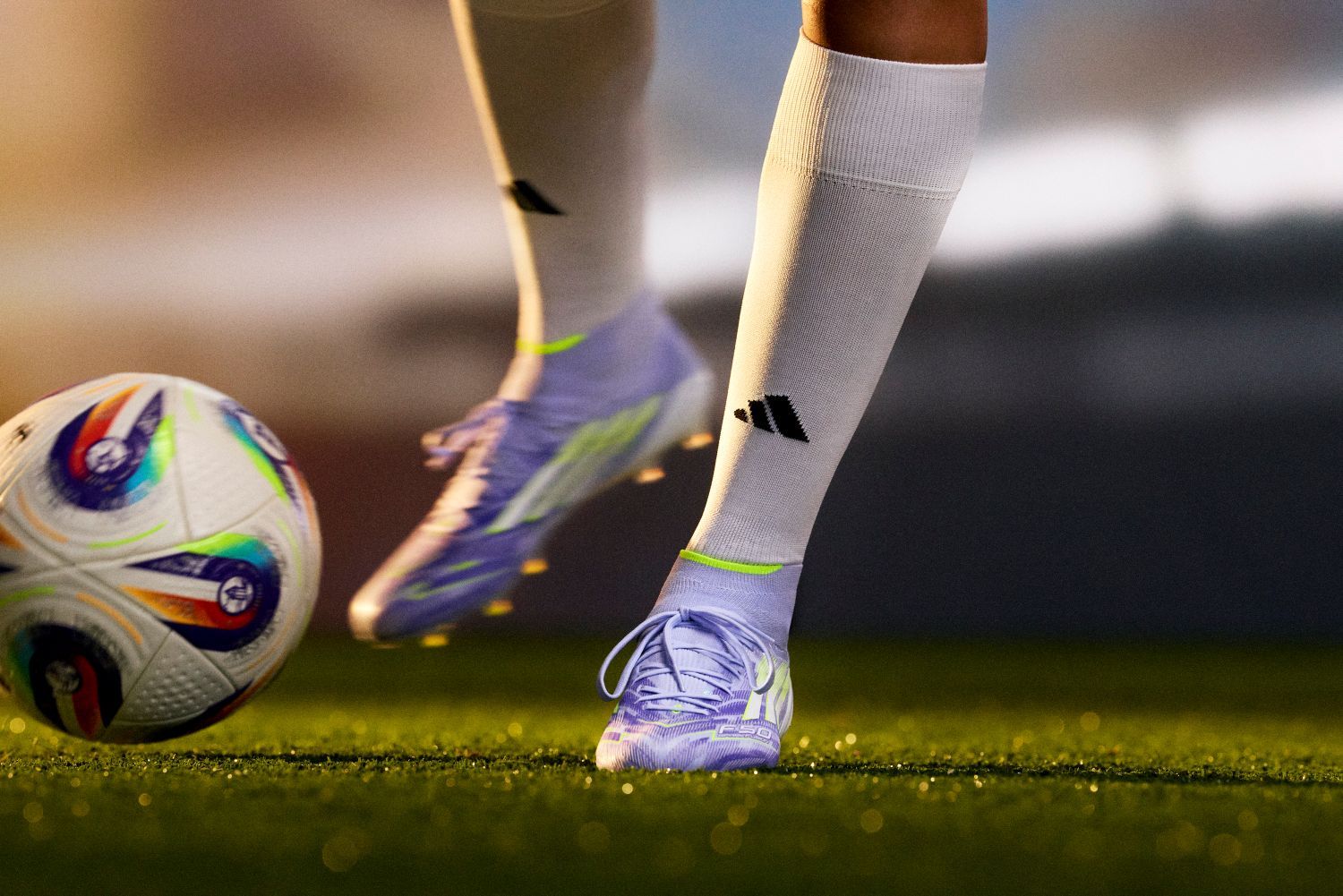 Close-up of a soccer player wearing purple adidas cleats and white socks, dribbling a colorful soccer ball on a grassy field.