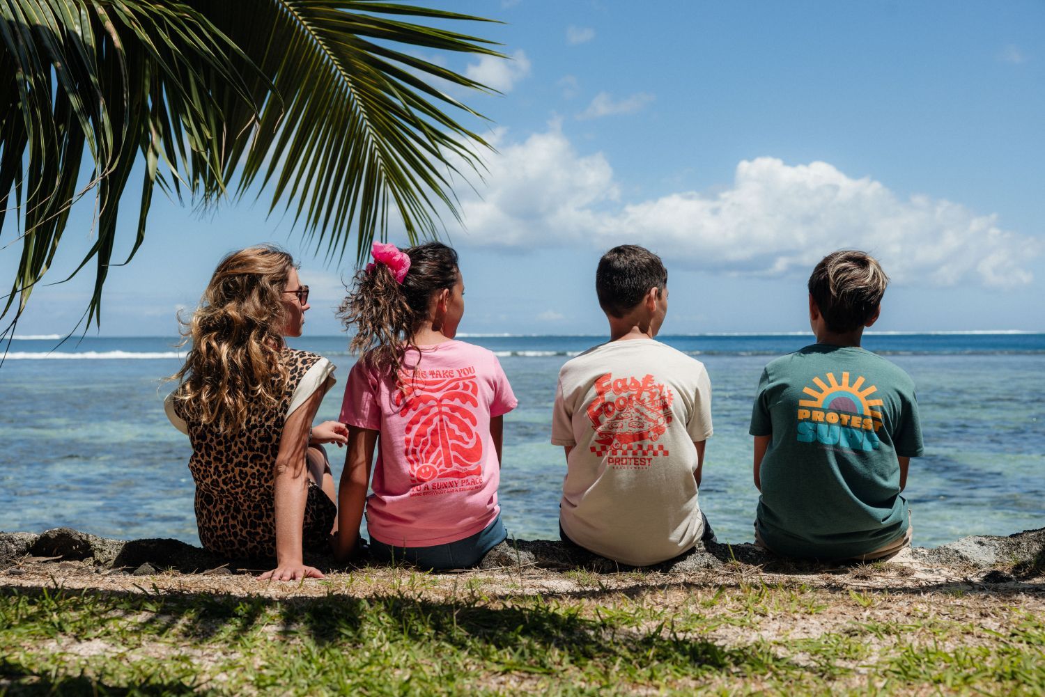 Vier kinderen van achteren gefotografeerd op een muurtje aan zee in Tahiti, dragend Protest T-shirts met kleurrijke backprints in roze, beige en groen onder een palmboom.