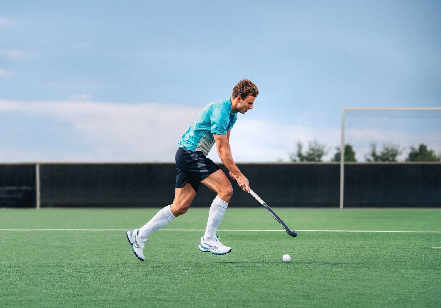 A man in a teal shirt plays field hockey on a green field, focusing on the ball.