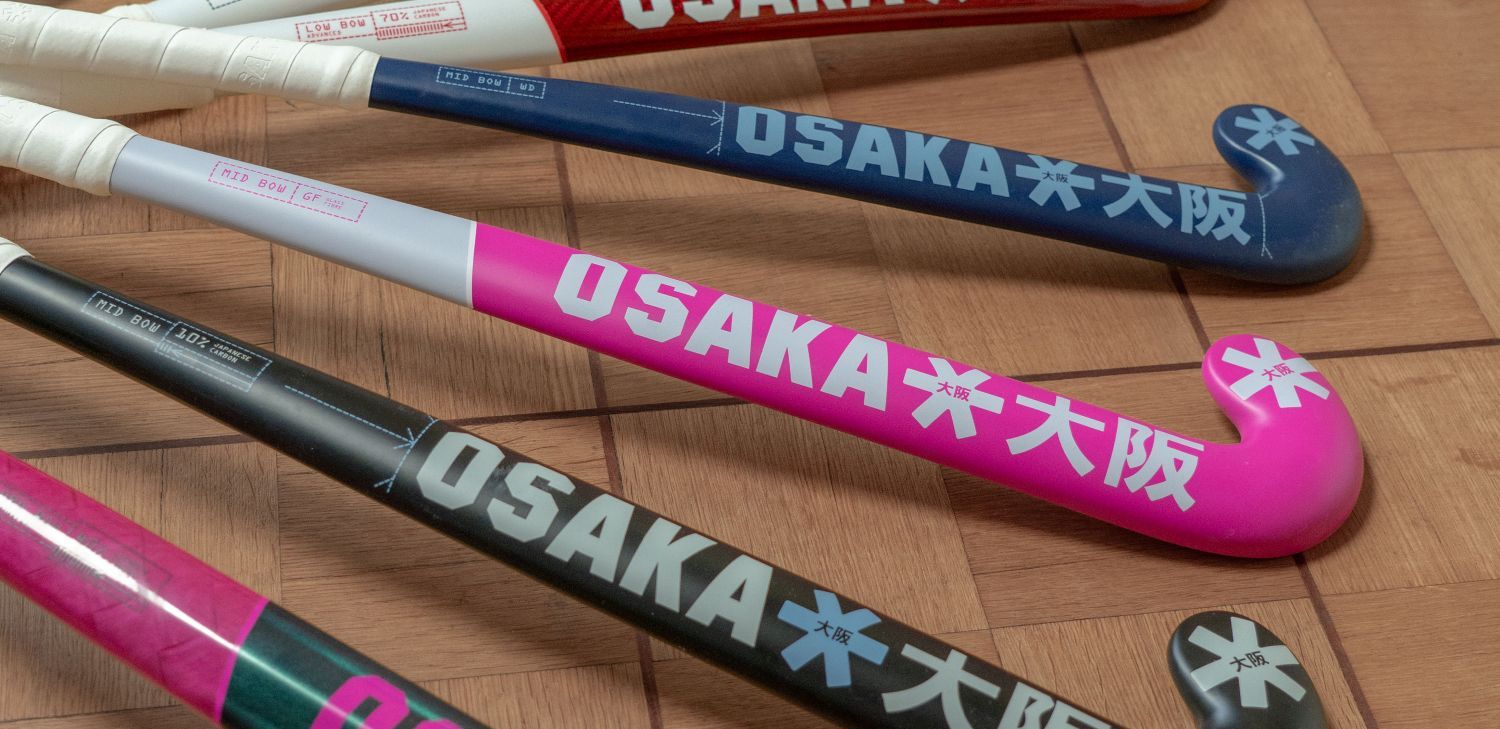 Welke hockeystick kiezen? Vind jouw perfecte match Colorful field hockey sticks with "OSAKA" branding, displayed on a wooden floor.