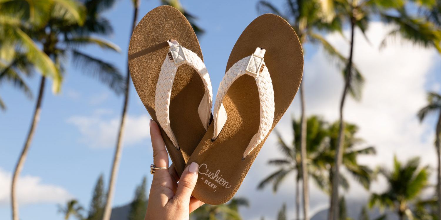 A hand holds up beige flip-flops with braided straps against a background of palm trees and a blue sky.