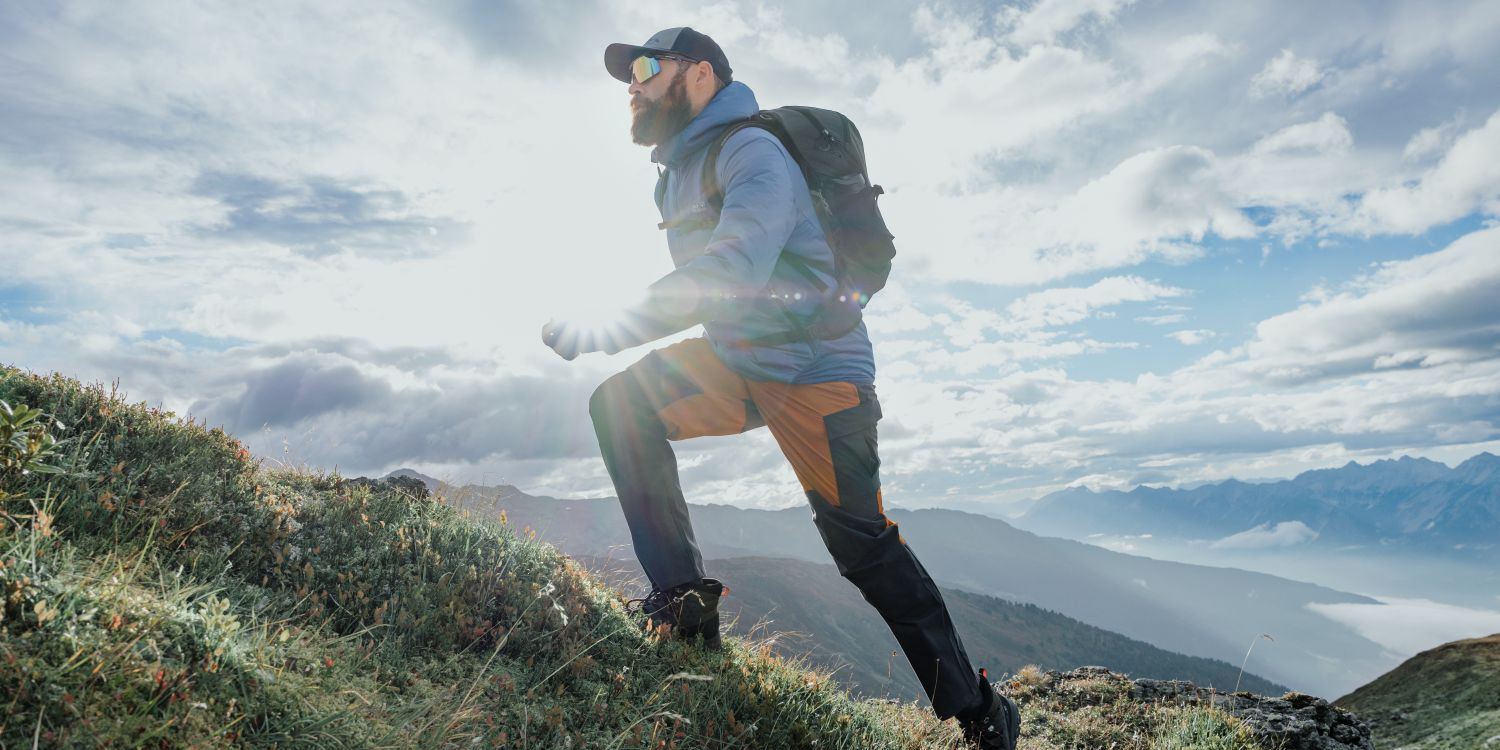 Man hiking uphill with a backpack in a mountainous landscape, sunlight shining behind him, under a partly cloudy sky.