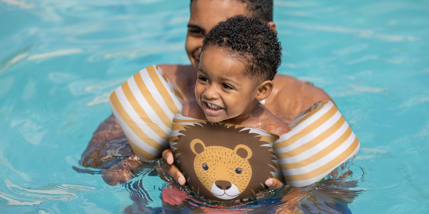 Child with lion-themed floaties smiles in pool, hugged by an adult. Bright, sunny day enhances the joyful atmosphere.