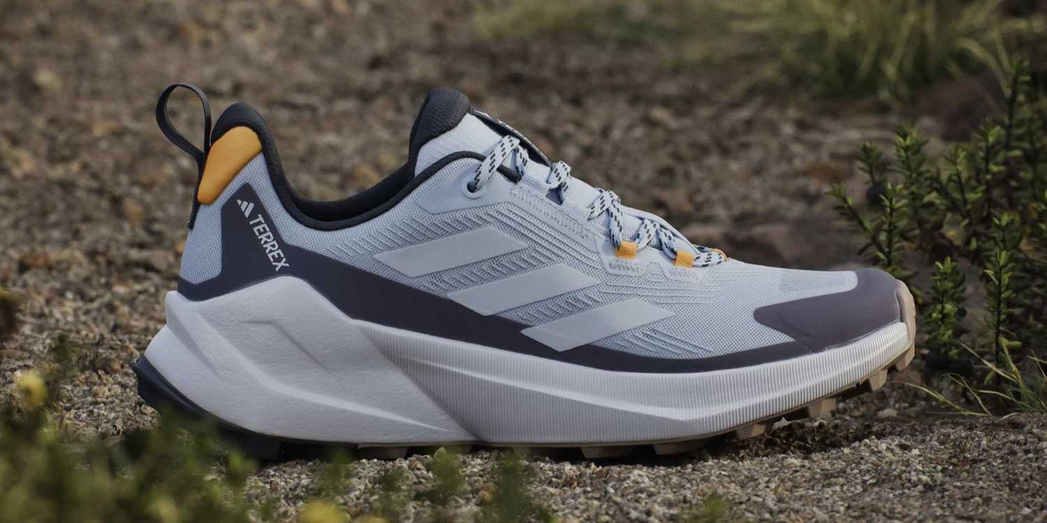 Light blue and gray trail running shoe with orange accents, set outdoors on a rocky and grassy terrain.
