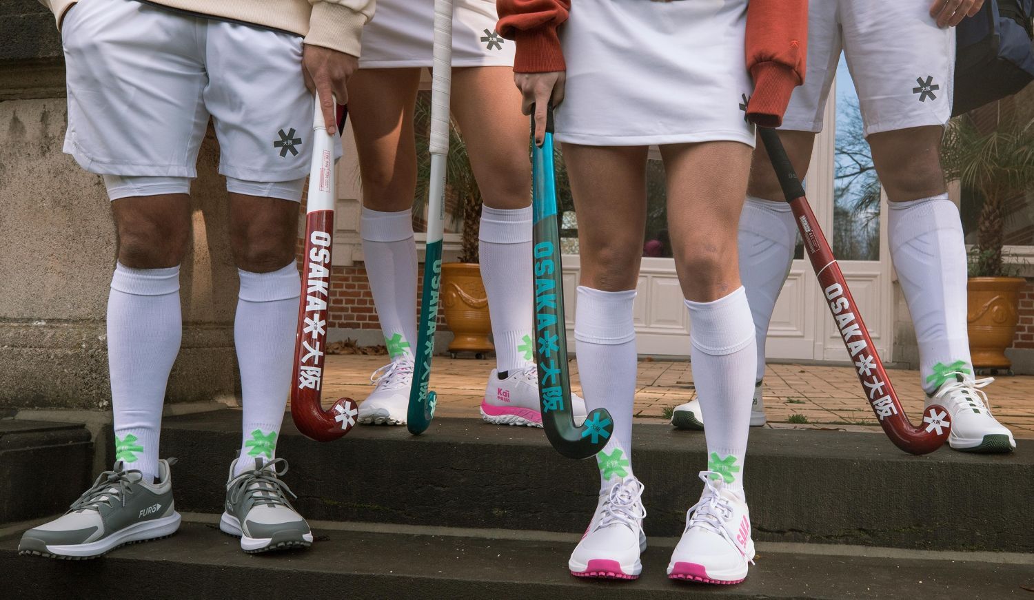 Hoe kies je de perfecte hockeyschoenen? De complete gids Group of field hockey players in white uniforms, holding colorful sticks, wearing matching socks and Osaka hockey shoes, standing on steps.