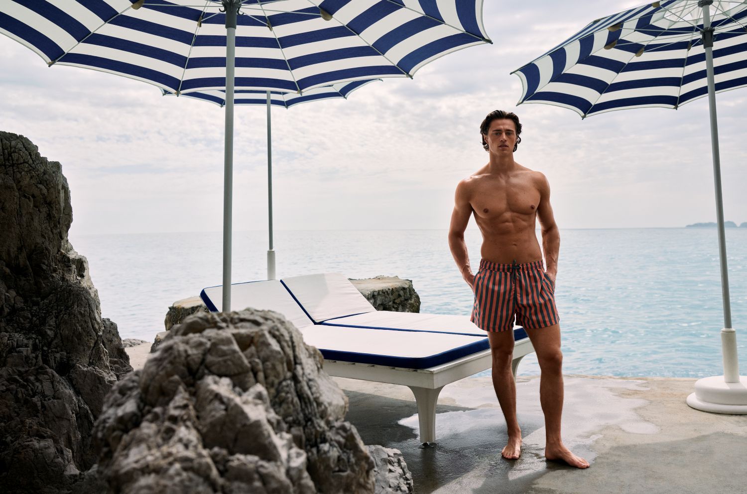 Gespierd model in gestreepte Björn Borg zwembroek bij ligbed en blauw-witte parasols aan de zee, Trend French Riviera.