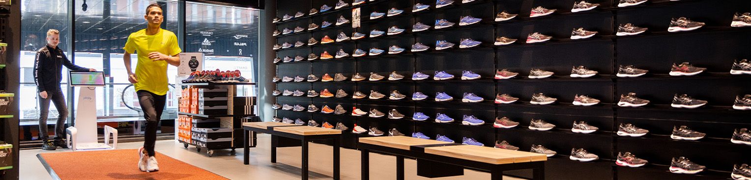 A person in a yellow shirt runs on an orange mat in a shoe store, surrounded by walls of various sneakers on display and another person observing.