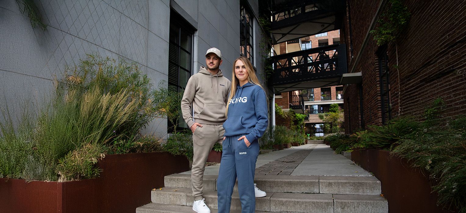 A man and a woman in casual hoodies pose confidently on an urban path flanked by greenery and modern buildings.