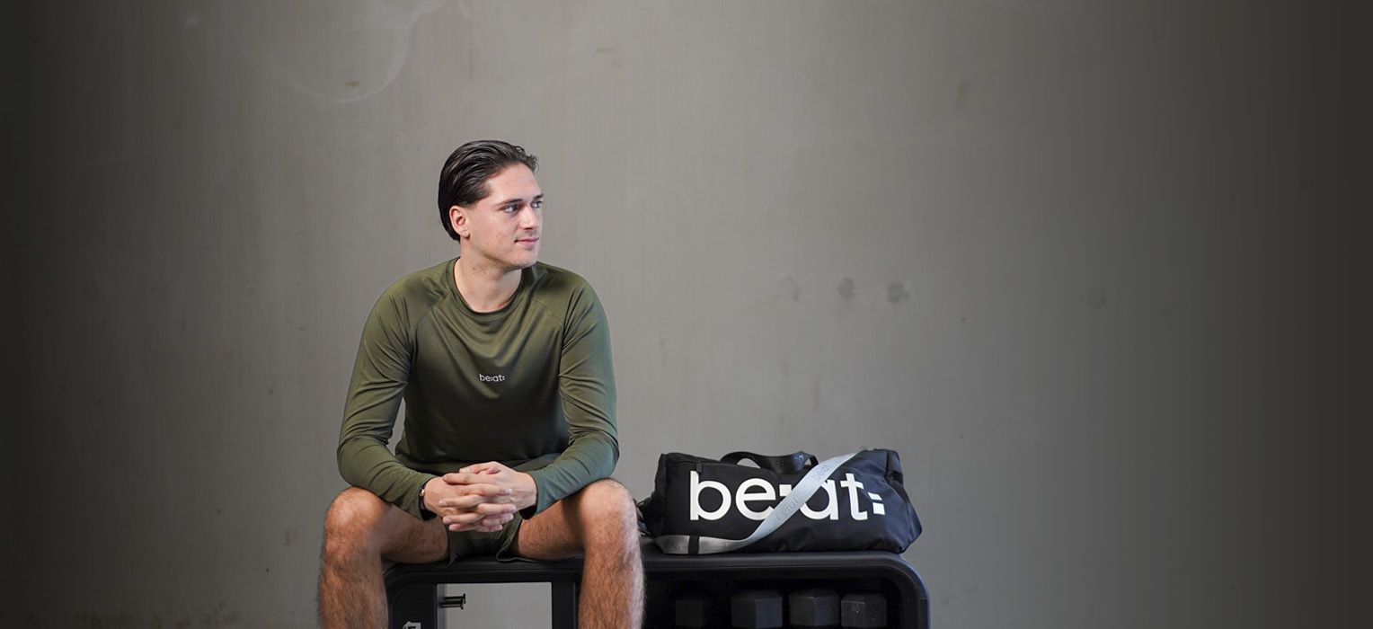 Man in a green long-sleeve shirt sits on a bench beside a black sports bag in a minimalistic setting.